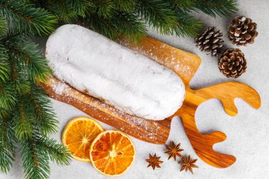 Klasik Noel Stollen, ahşap tahtada kurutulmuş portakallar, çam dalları ve beyaz desenli arka planda koniler ile pudra şekerli kremayla kaplıdır. Geleneksel bayram yemeği, tatlı meyveli ekmek.