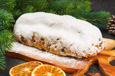 Klasik Noel Stollen, ahşap tahtada kurutulmuş portakallar, siyah desenli yüzey üzerinde çam dalları ve koniler ile pudra şekerli kremayla kaplıdır. Geleneksel bayram yemeği, tatlı meyveli ekmek.