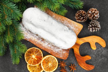 Klasik Noel Stollen pudra şekerli kremayla kaplanmış tahta tahtada kurutulmuş portakallar, siyah desenli yüzey üzerindeki çam dalları ve koniler, üst manzara. Geleneksel bayram yemeği, tatlı meyveli ekmek.