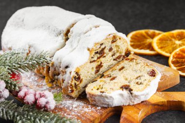 Geleneksel Noel süslemesi, ahşap tahtada dilimlenmiş tatlı meyve somunu, siyah desenli arka planda toz şekeri. Baharatlar, kurutulmuş meyveler ve şenlikli dekorasyonlar kış tatili havası yaratır.