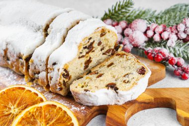 Geleneksel Noel süslemesi, ahşap tahtada dilimlenmiş tatlı meyve somunu, beyaz desenli arka planda pudra şekeri ile tozlanmış. Baharatlar, kurutulmuş meyveler ve şenlikli dekorasyonlar kış tatili havası yaratır.