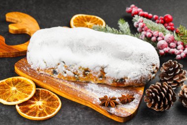 Klasik Noel Stollen, ahşap tahtada kurutulmuş portakallar, siyah desenli yüzey üzerinde çam dalları ve koniler ile pudra şekerli kremayla kaplıdır. Geleneksel bayram yemeği, tatlı meyveli ekmek.