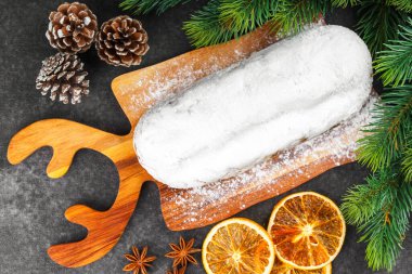 Klasik Noel Stollen pudra şekerli kremayla kaplanmış tahta tahtada kurutulmuş portakallar, siyah desenli yüzey üzerindeki çam dalları ve koniler, üst manzara. Geleneksel bayram yemeği, tatlı meyveli ekmek.