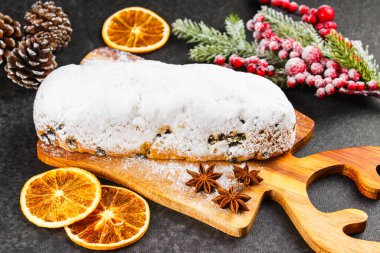 Klasik Noel Stollen, ahşap tahtada kurutulmuş portakallar, siyah desenli yüzey üzerinde çam dalları ve koniler ile pudra şekerli kremayla kaplıdır. Geleneksel bayram yemeği, tatlı meyveli ekmek.