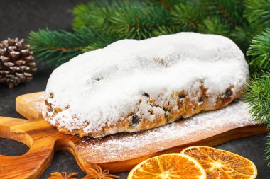 Klasik Noel Stollen, ahşap tahtada kurutulmuş portakallar, siyah desenli yüzey üzerinde çam dalları ve koniler ile pudra şekerli kremayla kaplıdır. Geleneksel bayram yemeği, tatlı meyveli ekmek.