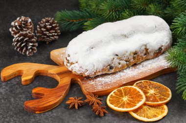 Klasik Noel Stollen, ahşap tahtada kurutulmuş portakallar, siyah desenli yüzey üzerinde çam dalları ve koniler ile pudra şekerli kremayla kaplıdır. Geleneksel bayram yemeği, tatlı meyveli ekmek.