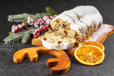 Geleneksel Noel süslemesi, ahşap tahtada dilimlenmiş tatlı meyve somunu, siyah desenli arka planda toz şekeri. Baharatlar, kurutulmuş meyveler ve şenlikli dekorasyonlar kış tatili havası yaratır.