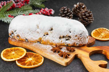Klasik Noel Stollen, ahşap tahtada kurutulmuş portakallar, siyah desenli yüzey üzerinde çam dalları ve koniler ile pudra şekerli kremayla kaplıdır. Geleneksel bayram yemeği, tatlı meyveli ekmek.
