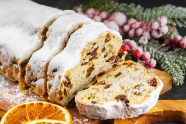 Geleneksel Noel süslemesi, ahşap tahtada dilimlenmiş tatlı meyve somunu, beyaz desenli arka planda pudra şekeri ile tozlanmış. Baharatlar, kurutulmuş meyveler ve şenlikli dekorasyonlar kış tatili havası yaratır.