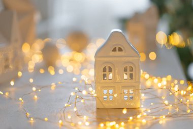 Beyaz seramik evi olan yumuşak bir Noel sahnesi, parlayan sıcak peri ışıkları ve tarafsız kış dekoru. Konforlu minimalist tatil arka planı mevsimlik tasarımlar için mükemmel.