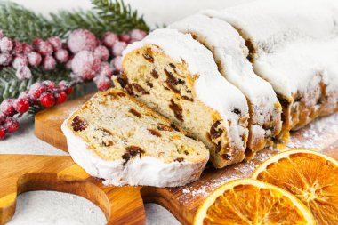 Geleneksel Noel süslemesi, ahşap tahtada dilimlenmiş tatlı meyve somunu, beyaz desenli arka planda pudra şekeri ile tozlanmış. Baharatlar, kurutulmuş meyveler ve şenlikli dekorasyonlar kış tatili havası yaratır.