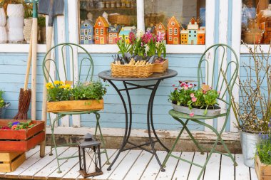 Çiçekçinin dış tarafı bahar çiçekleri, fidanlar, kır sandalyeleri ve masa ile süslenmiş. Pastel renklere sahip rahat bahçe atmosferi, doğal ışık ve el yapımı dekor, mevsimlik perakende, yaşam tarzı konsepti