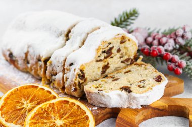 Geleneksel Noel süslemesi, ahşap tahtada dilimlenmiş tatlı meyve somunu, beyaz desenli arka planda pudra şekeri ile tozlanmış. Baharatlar, kurutulmuş meyveler ve şenlikli dekorasyonlar kış tatili havası yaratır.