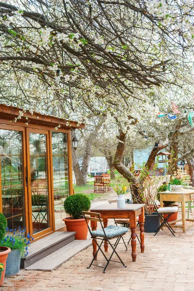 Tahta masa, sandalyeler ve çiçek açan ağaçlarla çevrili taze çiçeklerle büyüleyici bir bahar bahçesi sahnesi. Samimi açık kafe atmosferi, kırsal mobilyaları ve doğal mevsimsel manzarası var.