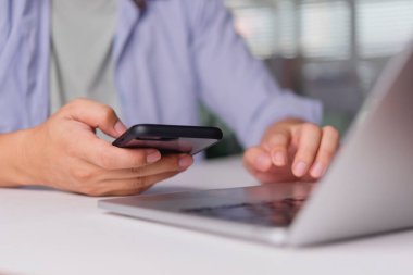 Freelancer çevrimiçi çalışmak ve iletişimi geliştirmek için ev bilgisayarında dizüstü bilgisayarda yazarken akıllı telefon, telefon ve cep telefonu kullanır; detaylı eller teknoloji ve verimliliği gösterir.