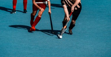 İki çim hokeyi oyuncusu, yapay çimlerde yoğun bir maç sırasında orta sahada top için dövüşüyor.