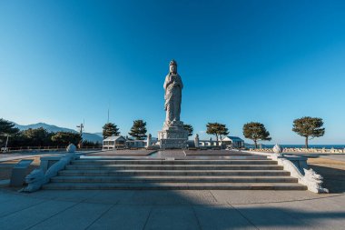 Granit Bodhisattva heykeli, Güney Kore 'deki Naksansa Tapınağı' nda, berrak mavi gökyüzünün altında, duaları ve sessiz yansımalarını çağıran basamakların üzerinde yükseliyor..