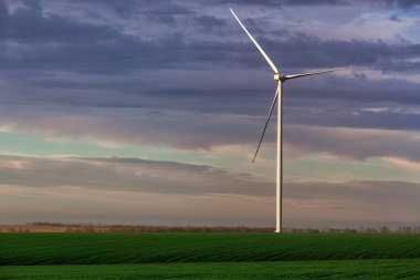 Arka plandaki yeşil tepelik tarımsal alanlarda rüzgar enerjisi jeneratörü. Bulutlar, sıfır emisyon. Açık hava çiftliğinde yenilenebilir eko enerji üretimi..