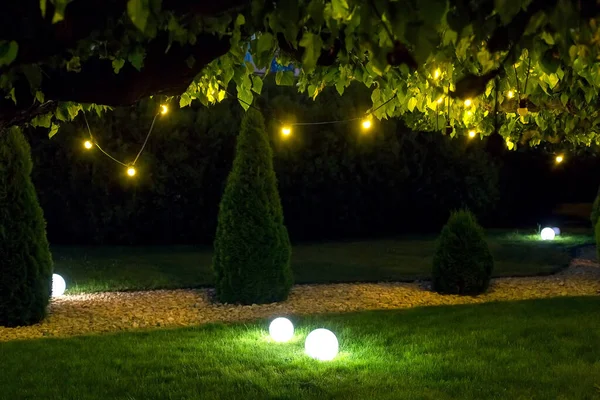 illumination park light garden with electric ground ball lantern with stone mulch and thuja bushes in outdoor landscaped park with garland of warm light bulbs, illuminate evening scene nobody.