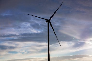 Demirden yapılma siluet rüzgar türbini, gün batımında bulutlu mavi gökyüzüne karşı bıçaklar, doğayı koruyan alternatif elektrik..