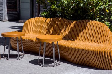 Üzerinde kahverengi ahşap tahtalar olan ahşap bir tezgah. Şehrin kaldırımında yuvarlak masalar var. Güneş ışığıyla aydınlatılmış..