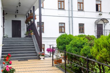 Verandanın önünde çiçekli saksılar. Tavan lambası beyaz bina, granit taşla kaplı ahşap korkuluklar. Arka bahçenin dış tarafında çalılar var..