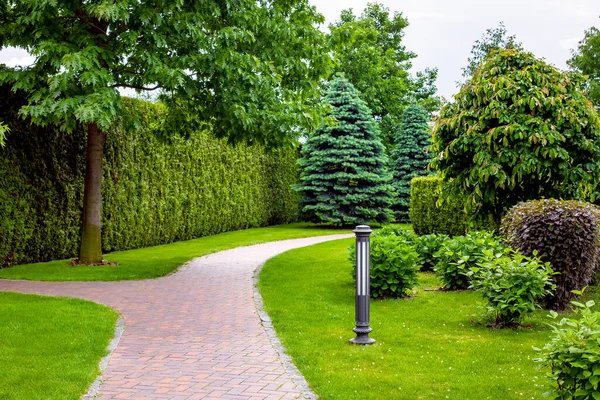 在公园里 石砖铺的人行道在常绿藤树篱和落叶树与铁皮花园灯笼的绿树中间 没有人 — 图库照片