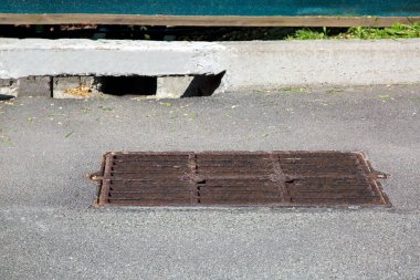 Asfalt bir yolda, dikdörtgen bir rögar deliğinde kanalizasyon ızgarası ve güneşli bir günde yol altyapısının kaldırımında bir fırtına hendeği kanalı..