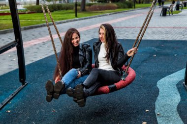 İki genç esmer kadın ve uzun saçlı kahverengi saçlı kadın, parkta deri ceketler, botlar ve kotlar içinde yuvarlak bir salıncakta oturan ve gülümseyen arkadaşlar..