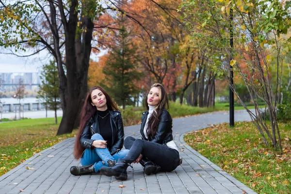 在秋天的公园里 两个年轻的女人坐在人行道上 靠着黄红的树叶 穿着皮夹克和牛仔裤 站在树后 她们的嘴唇在空中亲吻着 — 图库照片
