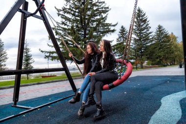 İki genç esmer kadın ve uzun saçlı kahverengi saçlı kadın, parkta deri ceketler, botlar ve kotlar içinde yuvarlak bir salıncakta oturan ve gülümseyen arkadaşlar..