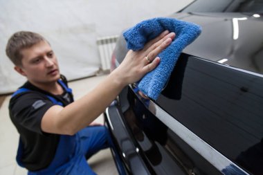 Bir erkek işçi siyah bir arabayı yıkar, suyu yumuşak bir bez ve mikrofiber ile siler, atölyede parlamak için yüzeyi temizler. Otomobil hizmet endüstrisi.