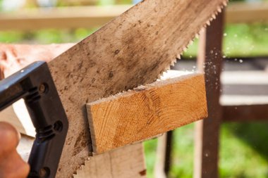 Marangozun elinde keskin dişleri olan siyah plastik saplı metal bir el testeresi kenarlarında küçük parçalar olan tahta kirişleri keserken. Bir yaz gününde. Kulübe inşaatı. 