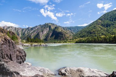Katun nehrinin kıyısında turkuaz kil ve kumlu bir plaj içeren su, dağların arka planında kayalık yeşil orman ve beyaz bulutlarla kaplı mavi gökyüzü..