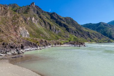 Katun nehrinin kıyısında turkuaz kil ve kumlu bir plaj içeren su, dağların arka planında kayalık yeşil orman ve beyaz bulutlarla kaplı mavi gökyüzü..