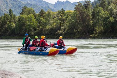 Altai, Rusya - 08.16.2020: Bir dağ nehrinde hızla kürek çeken mavi şişme bir teknede ekipman, can yelekleri ve sarı miğferler içinde rafting yapan insanlardan oluşan bir ekip. 