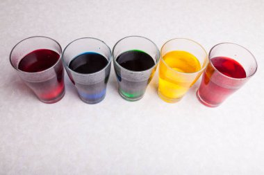 Beyaz bir mutfak masasında şeffaf bardaklar. İçi kırmızı mavi yeşil sarı, sulandırılmış gıda boyalarıyla dolu. Paskalya 'dan önce yumurtaları boyamak için hazırlanmış..