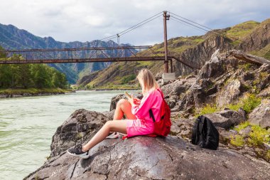 Pembe takım elbiseli genç, sarışın bir kadın Altai Dağları 'ndaki nehrin kenarındaki büyük bir taşın üzerinde oturuyor ve eski bir demir köprünün arka planındaki telefona bakıyor..