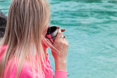 Pembe elbiseli, genç, sarışın bir kız Altai Dağları 'ndaki nehrin kenarındaki büyük bir taşın üzerine oturur ve dinlenirken demir bardaktan sıcak çay içer.