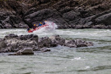 Turuncu can yelekleriyle rafting yapan bir grup insan bir dağ nehri boyunca mavi ve sarı renkli lastik bir botta ormanı olan kayalık bir sahilin arka planına karşı rafting yapıyorlar..