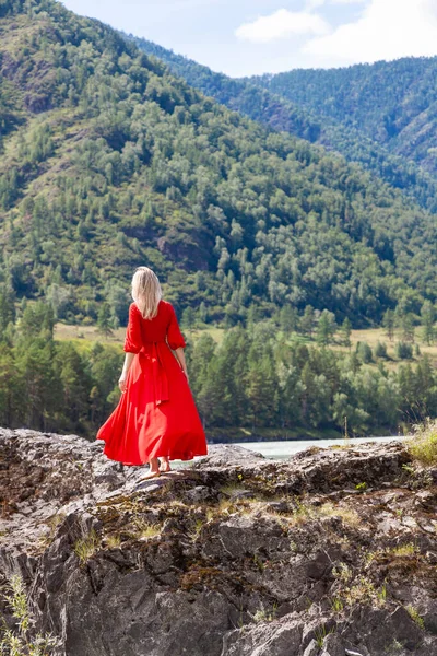 view from the back of Blonde woman in red dress fluttering in the wind stands on the rock stones on banks of Katun river against background of mountains on summer. WAiting for wonder. Assol.