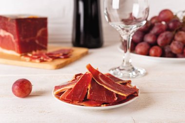 Kesme tahtasındaki büyük jamon parçasına karşı dilimlenmiş bir tabak, bir demet pembe üzüm, şarap şişesi ve beyaz ahşap masanın üzerinde cam. Lezzetli İspanyol jamonu Serrano. Ön görünüm.