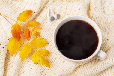 Büyük beyaz kupa sıcak kahve, kulaklık ve bej örülmüş atkıda sarı sonbahar yaprağı. Sıcak sonbahar konsepti. Üst görünüm.