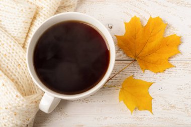 Beyaz ahşap kırsal yüzey üzerine bej örülmüş atkı yanında büyük beyaz fincanlı, dumanı tüten kahve ve iki sarı yaprak. Sıcak sonbahar konsepti. Üst görünüm. 