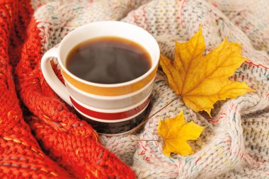 Kırmızı bej örgü eşarba sarılı çizgili dumanı tüten kahve kupası ve iki kuru sarı sonbahar yaprağı. Samimi düşüş konsepti. Üst görünüm.