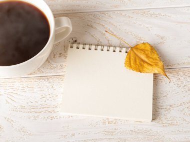 Açık ilkbahar not defterinde parlak sarı sonbahar yaprağı beyaz ahşap masanın üzerinde dumanı tüten bir fincan sıcak kahvenin yanında. Kopyalama alanı için boş sayfa. Üst görünüm. 