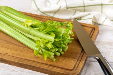 Bir demet taze yeşil kereviz sapı ve beyaz ahşap masanın üzerinde kahverengi bir kesme tahtasının üzerinde keskin şef bıçağı. Doğal antioksidan, sağlıklı beslenme ve kilo kaybı içeriği. Evde yemek pişir. Ön görünüm.