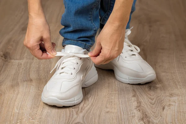 Caucasian woman hands tie elastic laces on chunky sole white sneakers shod on her feet on the brown floor. Pair of new comfortable shoes for active lifestyle, everyday life and sports. Low angle view.