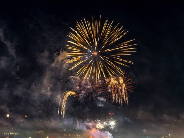 Karanlık gökyüzünde parlak havai fişek ışıkları. Küçük bir şehir gece vakti renkli havai fişeklerle kutlama yapıyor. Havai fişek bulutu gökyüzünde duman çıkarıyor. Gece açık hava sahnesi. Bayram havası ve bayram günleri. Gerçek hayat..