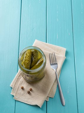 Cam bir kavanozda çıtır çıtır mısır turşusu ve mavi ahşap masa üzerinde çatal. Dereotu ve hardal tohumlarıyla marine edilmiş yeşil turşular. Lezzetli bebek turşusu. Lezzetli konserve sebzeler. Üst görünüm.
